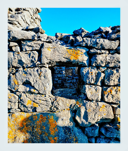 This photo shows a section of a weathered stone wall against a bright blue sky.  The wall is made of rough, irregularly shaped stones, many showing signs of age and discoloration.  Patches of orange lichen are visible on some of the stones.  A small, dark opening, a window, is visible in the center of the wall.