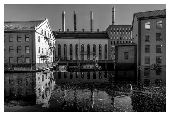 Ein industrielles Gebäudeensemble spiegelt sich in ruhigem Wasser, während hohe Schornsteine in den Himmel ragen und eine nostalgische Atmosphäre erzeugen.