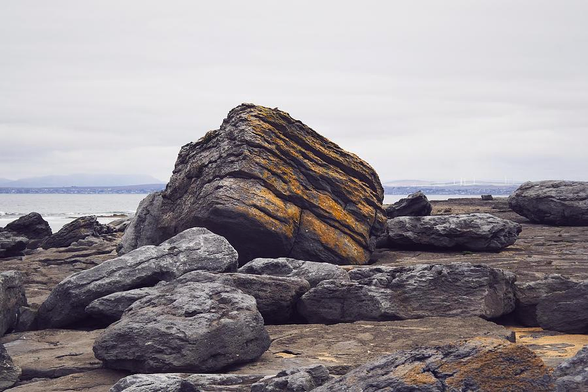 Large limestone boulders, with one particularly large one in part covered by rust-coloured lichens, lay strewn on limestone bedrock. Galway Bay / the Atlantic and the Connemara shore can be seen in the background.