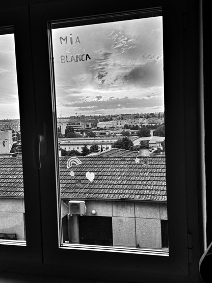 Una ventana muestra una vista en blanco y negro de una ciudad. La vista abarca tejados de casas con tejas, y al fondo se ve una ciudad con edificios de varios pisos bajo un cielo nublado. En el tejado más cercano a la ventana hay pequeños adornos en forma de corazón y arcoíris.  Un aparato de aire acondicionado se encuentra en la parte inferior de la imagen, en el tejado de un edificio.