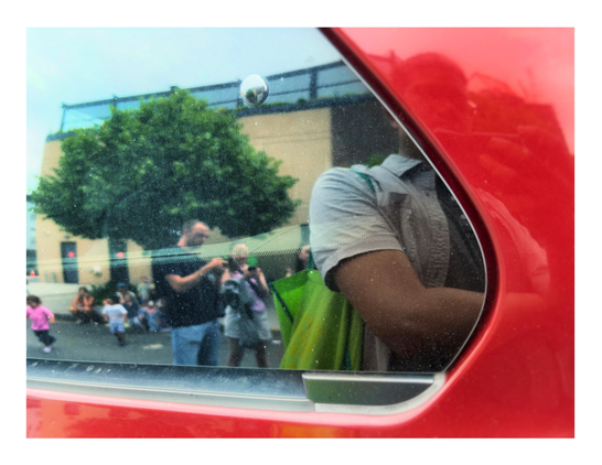 An outdoor city scene is reflected in the window of a parked red car. The reflection starts from left frame and takes up about 2/3rds of the image, bordered by the window’s curved diagonal edges. The car door’s glossy red surface serves as a frame at bottom and right. The reflection shows adults standing in the street while checking their phones, small children running around, a sidewalk tree, and a low-rise apartment building. I can be seen in the foreground of the reflection —  my arm and green tote bag appear in the window panel, while my face and my hand holding the iPhone are blurred by the red doorframe.
