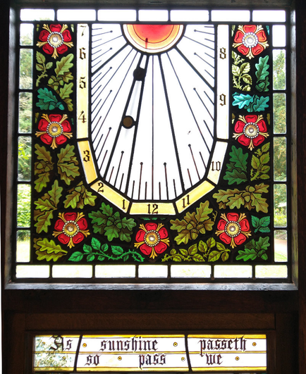 A colourful stained glass window with a sundial surrounded by red roses and green leaves.
The text below reads ‘As sunshine passeth so pass we’.
