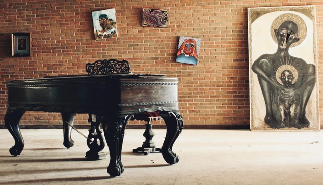 An ornate grand piano sitting in a room, with a brick wall behind it. A few haphazard pictures hang on the wall behind it and a large one is its to the right. This was taken in the lobby area of an abandoned university.