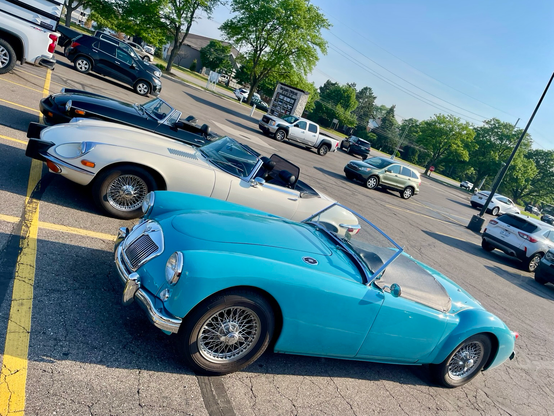 Photograph of three classic British sports cars in a parking lot with modern cars in the background. A blue MGA is in front with a white Jaguar E-Type behind and a black MGB behind that. The photograph tilts to the lower right.