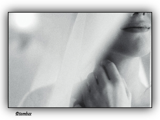 A black-and-white, blurred portrait of a young woman captured in an unconventional, fragmented composition. Only the lower part of her face is visible—sensual lips and a hand gently touching her neck. Part of her face is hidden behind a delicate, translucent curtain, creating an intimate, atmospheric mood. The soft blur enhances the sense of transience and mystery. Light falls gently, emphasizing the subtlety of the frame. The composition draws attention to its details and suggestions, leaving the rest to the viewer’s imagination.