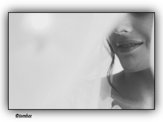 A black-and-white, blurred portrait of a young woman captured in an unconventional, fragmented composition. This time, not only are her lips visible, but also part of her nose—her lips slightly parted, adding subtle tension and expression to the image. A delicate, translucent curtain still conceals part of her face, creating an intimate and enigmatic mood. The soft blur deepens the sense of transience and mystery. Gentle light envelops the frame, emphasizing its sensual and atmospheric character.