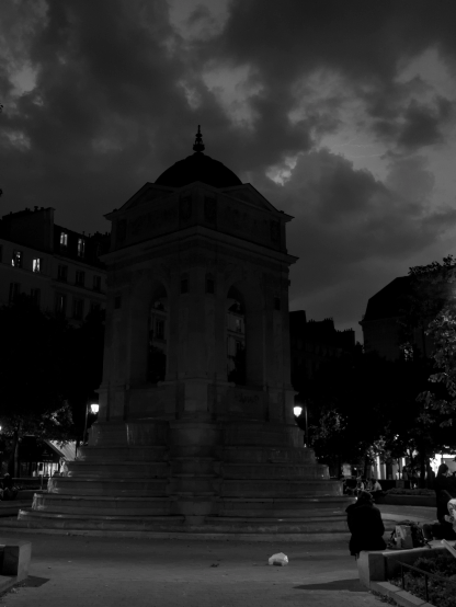 Monochrome photography of a kiosk-shaped fountain in a Parisian square, under a covered sky and a bit of lightning visible between the clouds .