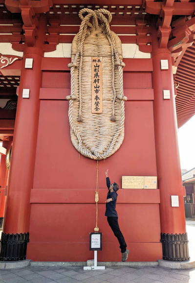 A giant waraji (straw sandal) hanging on the red painted wooden gate at Sensoji temple in Asakusa, Tokyo. A person is jumping and feet are off the ground as he is reaching out to try and touch the (bottom of the) waraji