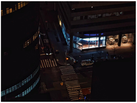 Nighttime shot from a window in a tall building looking down at a street corner with the bottom corner of the building diagonally opposite lit up. 
Zebra crossings are visible and a curved building is to the left with a few floors with light in the windows