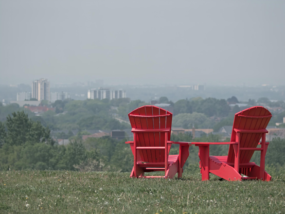 Two red Adirondack chairs positioned on a grassy hill overlooking a cityscape. The chairs are placed side by side, facing away from the viewer, and are accompanied by a small table between them. The chairs are bright red and have a classic design, with wide armrests and a sloping back