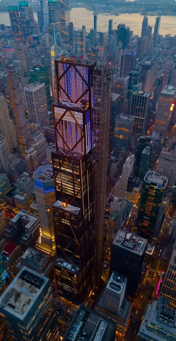 The image depicts a cityscape at dusk or dawn, characterized by numerous high-rise buildings. The central focus is on a tall skyscraper with a distinctive grid-like facade, surrounded by other illuminated buildings. The city appears densely populated with buildings of various architectural styles and heights. The background shows a body of water, likely a river, with more skyscrapers lining the horizon. The overall ambiance suggests a bustling metropolitan area, possibly New York City.