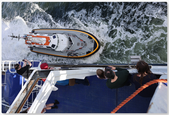Ein kleineres Boot nähert sich der großen Fähre von Amsterdam nach Newcastle. Zwei Männer strecken ihre Arme Richtung Schiff aus, um eine Person an Bord zu holen. Menschen fotografieren das Manöver vom Deck aus.

A small boat approaches the large ferry from Amsterdam to Newcastle. Two men stretch out their arms towards the ship to get a person on board. People photograph the manoeuvre from the deck.