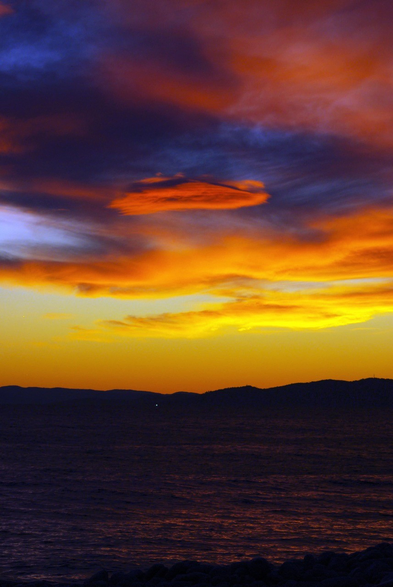 Photo au format vertical d’une plage juste après que le soleil a disparu derrière l’horizon. La mer est d’un beau violet sombre avec des reflets rose-orangés, et est bordée dans le fond par des collines basses qui se découpent en noir devant le ciel jaune orangé. Au dessus, des nuages comme peints par la lumière, avec un dégradé de rose, violet sombre, orange et jaune.