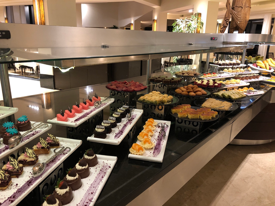 Das Bild zeigt eine große, reichhaltig bestückte Dessert-Auslage in einem Hotel oder Restaurant. Auf mehreren Ebenen und Tabletts sind zahlreiche verschiedene Kuchen, Törtchen, Gebäckstücke und andere Süßwaren präsentiert. Die Auswahl reicht von kleinen, feinen Törtchen über orientalisch anmutendes Gebäck bis hin zu größeren Kuchenstücken. Die Anordnung ist ansprechend und die Desserts sehen appetitlich aus.  Im Hintergrund ist ein Teil des Restaurants zu sehen.