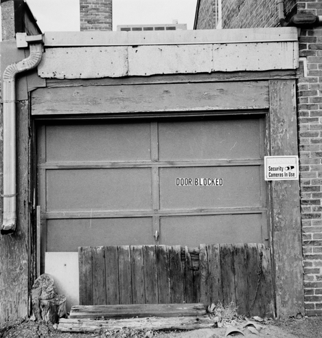 Photo, black and white, square format. Flat view of a back alley garage that has been haphazardly boarded at the bottom with recycled boards, loose bits of wood, and a single chunk of tree stump. Letter stickers are affixed, and read 'door blocked' in all capital letters. A small metal sign is hung to the right of the door frame, alerting passers-by that security cameras are in use.