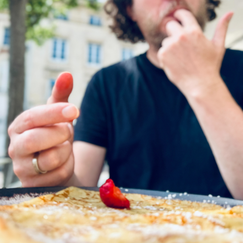 Ein Mann sitzt im Freien an einem Tisch und isst eine Crêpe. Im Vordergrund ist die Crêpe mit einer roten Erdbeere und Puderzucker zu sehen. Der Mann trägt ein schwarzes T-Shirt und leckt sich den Finger ab. Der Hintergrund ist unscharf, zeigt jedoch Gebäude und Bäume.