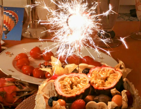 Zwei Teller. Auf den vorderen Teller eine Torte mit verschiedenen Früchten obenauf, ua eine aufgeschnittene Passionsfrucht mit den Fruchtseiten nach oben. Auf dem hinteren Teller liegen kleine rote Tomaten mit Zahnstochern zu Herzchen zusammengesteckt. Eine brennende Wunderkerze steckt in der Torte und dominiert das Bild mit brennenden Sternchen.