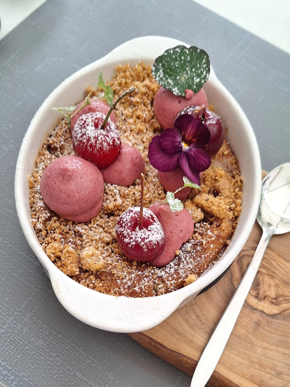 The image features a beautifully presented dessert in a white oval dish. The dessert consists of a crumbly, golden-brown base, likely a crumble or streusel topping, dusted with powdered sugar. On top of the crumble, there are several pink scoops of what appears to be a berry-flavored sorbet or ice cream, garnished with powdered sugar. Additionally, there are whole cherries with stems, some of which are dusted with powdered sugar, adding a vibrant red color to the dish. A delicate purple flower, possibly a pansy, and a green leaf are used as garnishes, adding a touch of elegance. The dish is placed on a wooden board, which rests on a gray textured surface, and a silver spoon is positioned to the right of the dish. The overall presentation is visually appealing and suggests a carefully crafted dessert.

Provided by @altbot, generated privately and locally using Ovis2-8B

🌱 Energy used: 0.227 Wh
