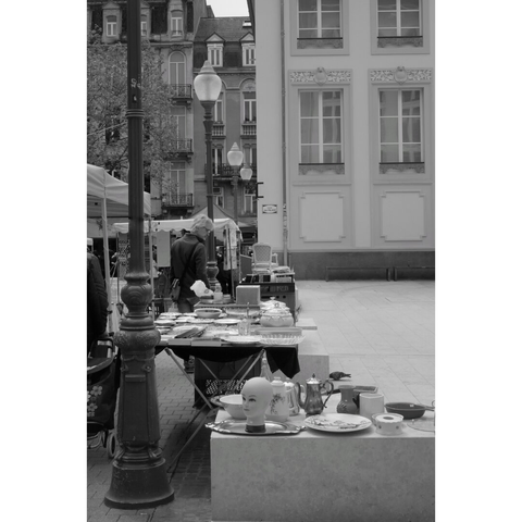 Black and white street photography taken in Luxembourg city. Every Saturday, on the place d'armes, in the center, a small flea market is organized with overpriced items, like old Villeroy plates or, on the front of the picture, the bald head of a big puppet. A man, maybe older than what is presented, is inspecting some objects in the background.