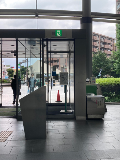A glass entrance to a building with an emergency exit sign above. Visible outside are pedestrians, a traffic cone, and greenery. An angular metal structure is in the foreground, with a bin beside it. The atmosphere is urban and slightly cloudy.
