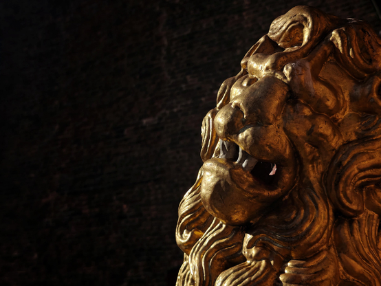 Golden lion sculpture photographed in sun light against a very dark background. The lion's head is shown in profile with mouth open, slightly turned towards the camera.