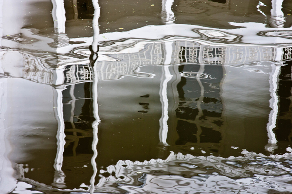 A reflection of the front of a stately, white Hague villa in a canal. You only see a terrace and some windows in the photo, upside down. The image in the water has become black and white. The poles of the terrace and the styles of the windows are a bit crooked by the wind. The windows look like dark holes. The whole thing gets the atmosphere of a lost place.