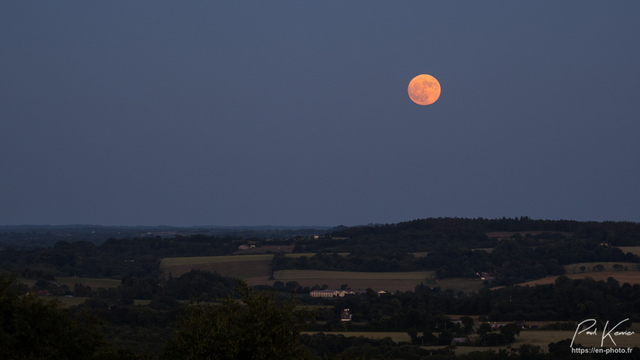 Photo en couleur, présentant la pleine lune des fraises, pas encore très haut dans le ciel, au-dessus d'une campagne vallonnée. comme la dénomination de cette lune semble l'indiquer, sa couleur, du moins quand elle est encore relativement proche de l'horizon, peut faire penser à la couleur du fruit.