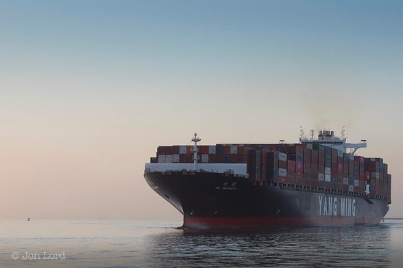 This Is A Colour Seascape Photo In Landscape Format. The Lower Ten Per Cent Of The Image Is Filled With A Body Of Smooth And Still Ocean. The Water Is Brighter On The Left Side From The Still Low Dawn Sun And Darker On The Right Side. Stretching From The Centre To The Right Margin Is A Large Container Ship That Fills A Quarter Of The Frame. The Ship Is Riding High In The Water With About Two Metres Of Red Anti-Fouling Paint Visible Above The Waterline. Perhaps The Ship Has De-Ballasted In Order To Enter The Harbour. As Is Common With Merchant Ships The Hull Is Black, While The Superstructure, Located About Three Quarters Of The Way Aft, Is White. The Shipping Line's Name: 'Yang Ming' Is In Large Bold White Letters On The Side Of The Hull While The Ships Name: 'YM Unanimity' Is In Smaller Letters Aft Of The Bow. Above The Deck Is Filled With Thousands Of Multi Coloured Shipping Containers, Mostly Forty Feet Long. The Ship Is Under Way From Right To Left But Moving So Very Slowly She Hardly Has Any Wake To Break The Calm Ocean Surface. Beyond The Ship Is A Crisp And Clear Horizon. Above Is An Orange / Golden Glow From The Low Rising Sun. The Clear And Cloudless Sky Graduates To Pale Blue Towards The Top Of The Image. 
The Location: The San Pedro Channel Outside The Port Of Los Angeles (2018).