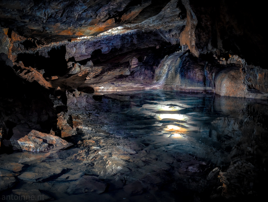 The interior of the Erdmannshöhle cave, dominated by a large, still body of water reflecting light from above. The cave walls and ceiling are rugged and irregular, showcasing various rock formations in shades of dark brown, orange, and hints of blue and purple. Along the edges of the water and on the cave walls, there are visible stalactites and stalagmites.
