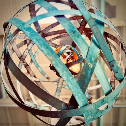 A loose, spherical chandelier comprised of turquoise, criss-crossing metal slats wrapped around a much smaller, similar sphere housing two Edison bulbs hangs from a tall, white ceiling in the distance.