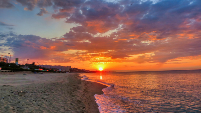 La imatge mostra una posta de sol impressionant sobre el mar. El cel està ple de colors taronja i vermell vibrants, amb núvols que semblen pintats amb pinzellades de foc. El sol està a prop de l'horitzó, reflectint la seva llum daurada sobre l'aigua. Una platja de sorra s'estén al llarg de la vora inferior de la imatge, amb una costa tranquil·la on onades suaus arriben a la riba. Al fons, es poden veure edificis i una mica de vegetació, donant un toc de civilització a l'escena. El conjunt crea un ambient serè i espectacular.