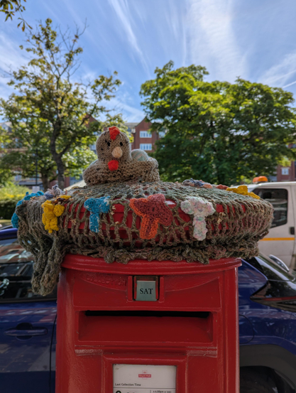 Red postbox on a street in Southport, UK covered on top with a yarn-stitched or crocheted chicken 🐔