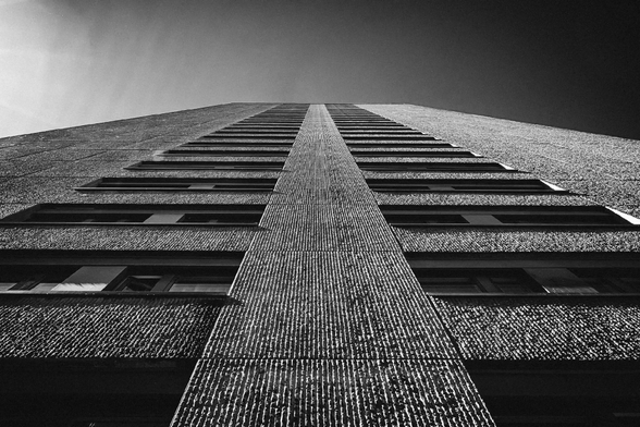 Dramatic low-angle view of a tall concrete building with rough-textured vertical panels and evenly spaced windows, captured in black and white. The image emphasizes the building’s height and symmetry as it stretches toward a gradient sky.