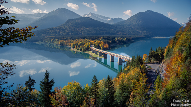 This stunning image captures a bridge over a lake with majestic mountains in the background. The scene is surrounded by lush green trees and bushes, creating an idyllic atmosphere. The sky is bright blue, adding to the beauty of this highland landscape. In the foreground, we can see a road leading up to the bridge which crosses over the body of water below. On either side of this road are tall trees with yellow and green leaves that stand out against their surroundings. Further away from us lies a mountain range covered in dense forest, providing an impressive backdrop for this tranquil setting. This picture perfectly encapsulates nature's beauty and serenity