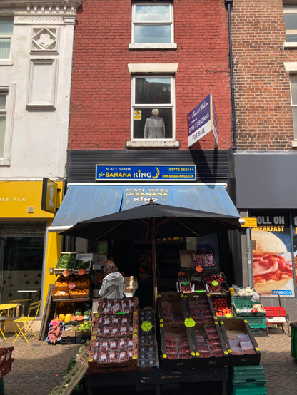 photo of a greengrocer with produce outside, shop sign "Matt Wade the banana king". in the first floor window, a lifesize cardboard cutout of King Charles looking out