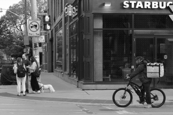 In the right foreground is a food courier on a bike, waiting for the light. At extreme left, are three people with a small dog, also waiting to cross. In the background at right, on the corner is a Starbucks. 