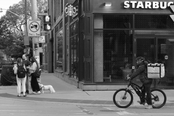 In the right foreground is a food courier on a bike, waiting for the light. At extreme left, are three people with a small dog, also waiting to cross. In the background at right, on the corner is a Starbucks.