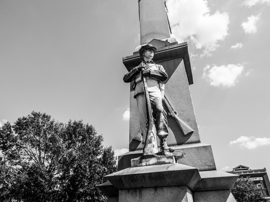 A black and white photograph of a Civil War memorial statue viewed from below against a cloudy sky. The bronze or stone figure depicts a uniformed soldier wearing a wide-brimmed hat and holding a rifle, standing at attention on a tall rectangular pedestal. The monument is surrounded by mature trees, and a historic building with classical architecture is visible in the background to the right. The low angle of the photograph emphasizes the statue's imposing presence and commemorative nature. The dramatic sky with scattered clouds creates a solemn, dignified atmosphere typical of war memorials.