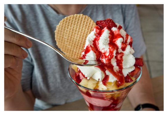 The picture shows a sundae being eaten by a person with a spoon. The glass contains various layers of pineapple chunks and strawberries. On top is a large portion of whipped cream decorated with strawberry sauce. On top of the cream is a fresh strawberry. Inside the whipped cream is a crispy, honeycomb-shaped round wafer cookie.