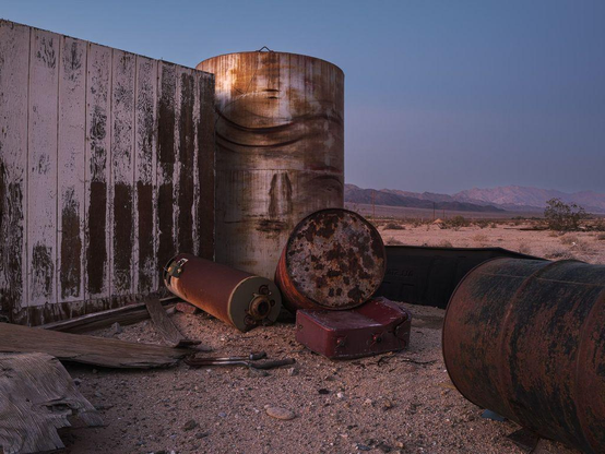 Several rusty barrels and worn-out shed in desert landscape with mountains in background, in after-sunset light.