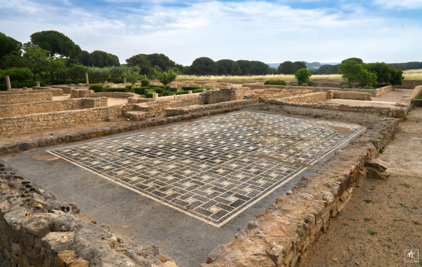 Color photo of a black and white patterned mosaic floor surrounded by the low ruined remnants of stone walls. In the background are the ruins of the buildings of an ancient Greek and Roman city with trees & fields beyond. 