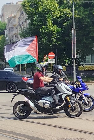 Photographie prise dans la rue à un carrefour.
À l'arrière à gauche, bâtiment et à droite arbre.
Au milieu drapeau palestinien fixé sur une mobilette qui passe.
Un homme à l'arrière fait le v de la victoire.
À l'avant, la rue avec les rails de tram.