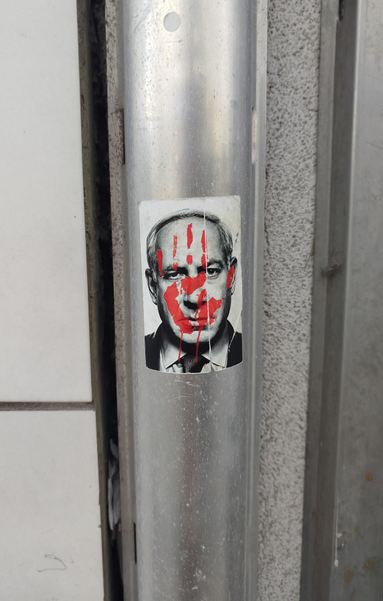 Stiker sur un poteau dans la rue représentant Netanyahou en noir et blanc avec sur son visage une empreinte de main gauche rouge.