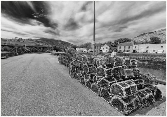 A black and white image of a harbor area featuring a stack of lobster traps on a paved road, with houses and hills visible in the background under a dramatic sky.
