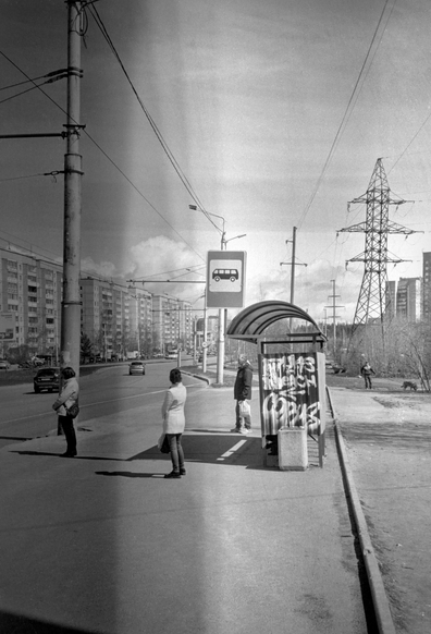 A monochrome photo shows people standing at a bus stop. Contrasting shadows fall from the people and from the bus stop.  The side of the bus stop is painted with graffiti. Behind the bus stop is a panorama