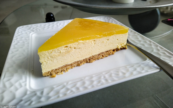 Auf einem weißen, quadratischen Kuchenteller liegt ein Stück Sanddorntorte, rechts daneben eine Kuchengabel.
Der Kuchen besteht aus drei Teilen: einem knusprigen braunen Boden, einer leichten Quark-Sahne-Füllung und einer gelben Schicht aus eingedicktem Sanddornsaft. /
A slice of sea buckthorn cake lies on a white, square cake plate, with a cake fork to the right. The cake consists of three parts: a crispy brown base, a light quark and cream filling, and a yellow layer of thickened sea buckthorn juice.
