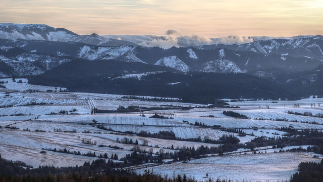 Late afternoon photo of a large valley with a mountain range on the opposite side in the distance. There are small hills on the floor of the valley, with fields and pastures covered by a thin layer of snow. The sky is partly cloudy and yellow from the sunlight.