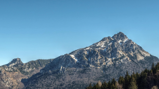 Photo of a mountain, back in the distance with a rocky, partially snow-covered top and forest growing on the lower parts, partially obscured by a forest of evergreen trees closer by.