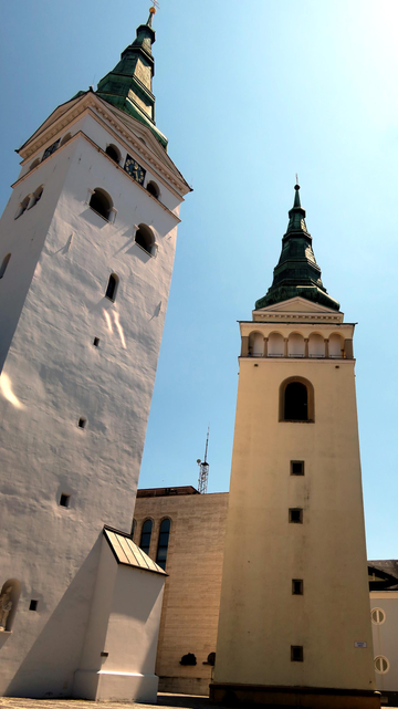 Photo of two high bell-towers with rectangular footprint standing on a small square. Both have spires on top covered by copper plates corroded green. There is a rectangular building in the background with marble facade. The sky is clear and blue.
