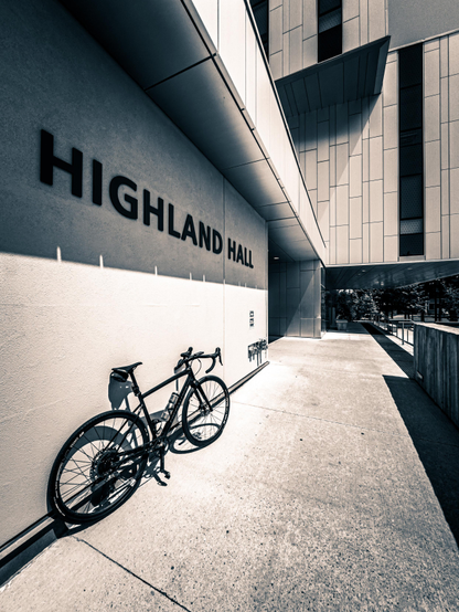 monochrome photo of a black specialized diverge gravel bike leaning against a concrete wall. the wall has the words “HIGHLAND HALL” written it. beyond the bike are portions of modern looking building exiting the frame towards the sky